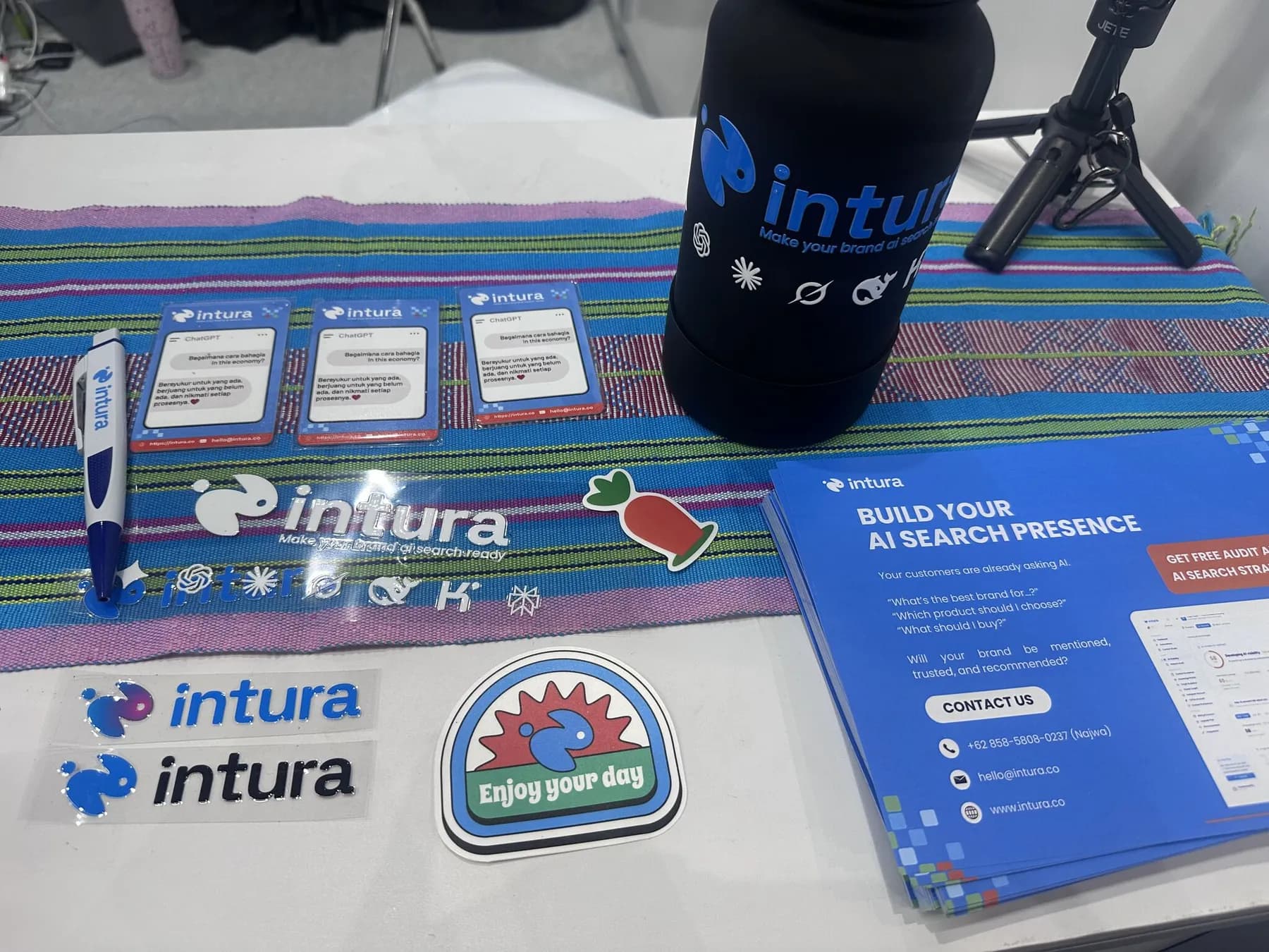 Merchandise booth Intura di INTI 2026 — stiker, pulpen, tumbler, dan flyer berjudul "Build your AI search presence"