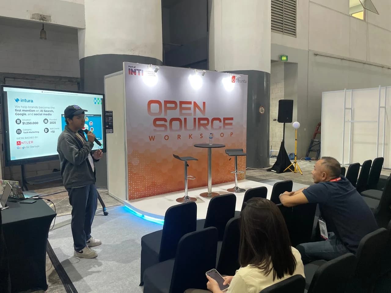 Intura tampil di panggung Open Source Workshop INTI 2026 — slide bertuliskan "We help brands become the first mention on AI Search, Google, and social media"
